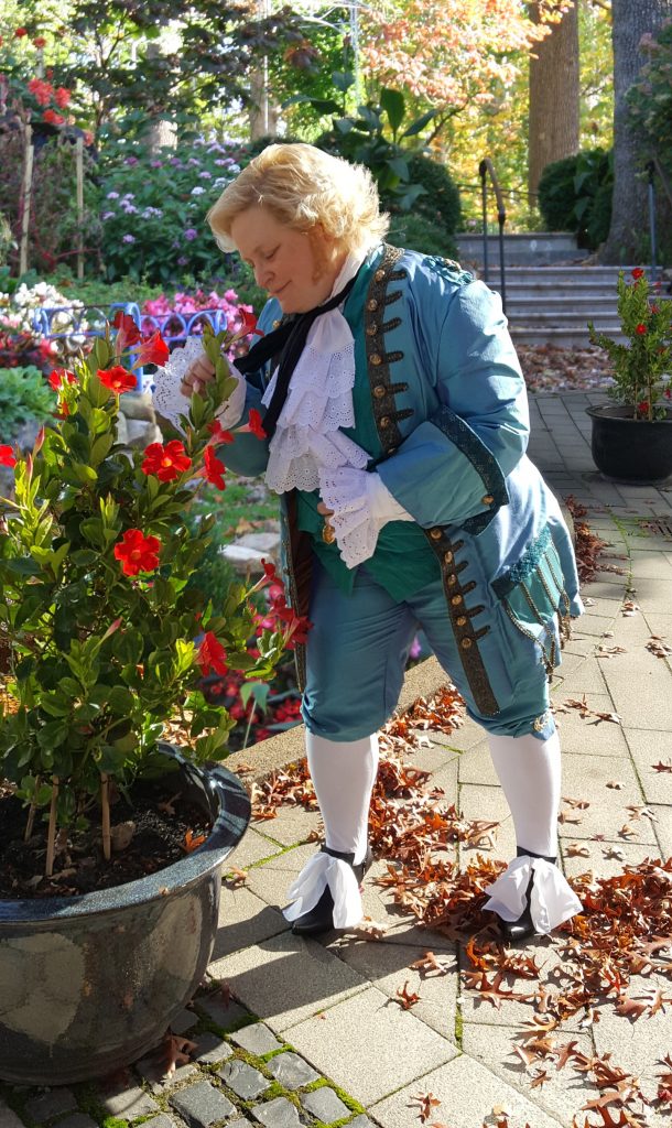 Mel in their self-sewn turquoise costume of Stede Bonnet. They lean over a potted flower to smell them.